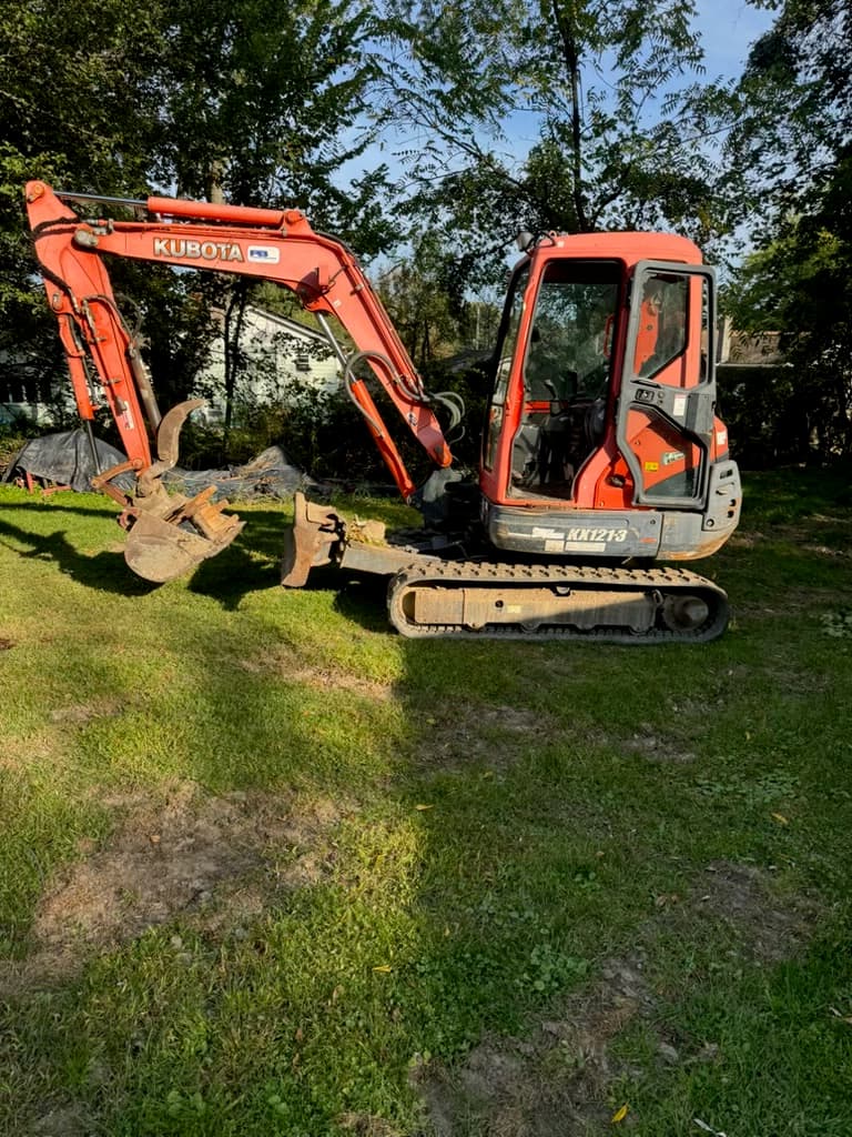 Orange Kubota KX121-3 mini excavator parked on a grassy lawn with trees in the background.