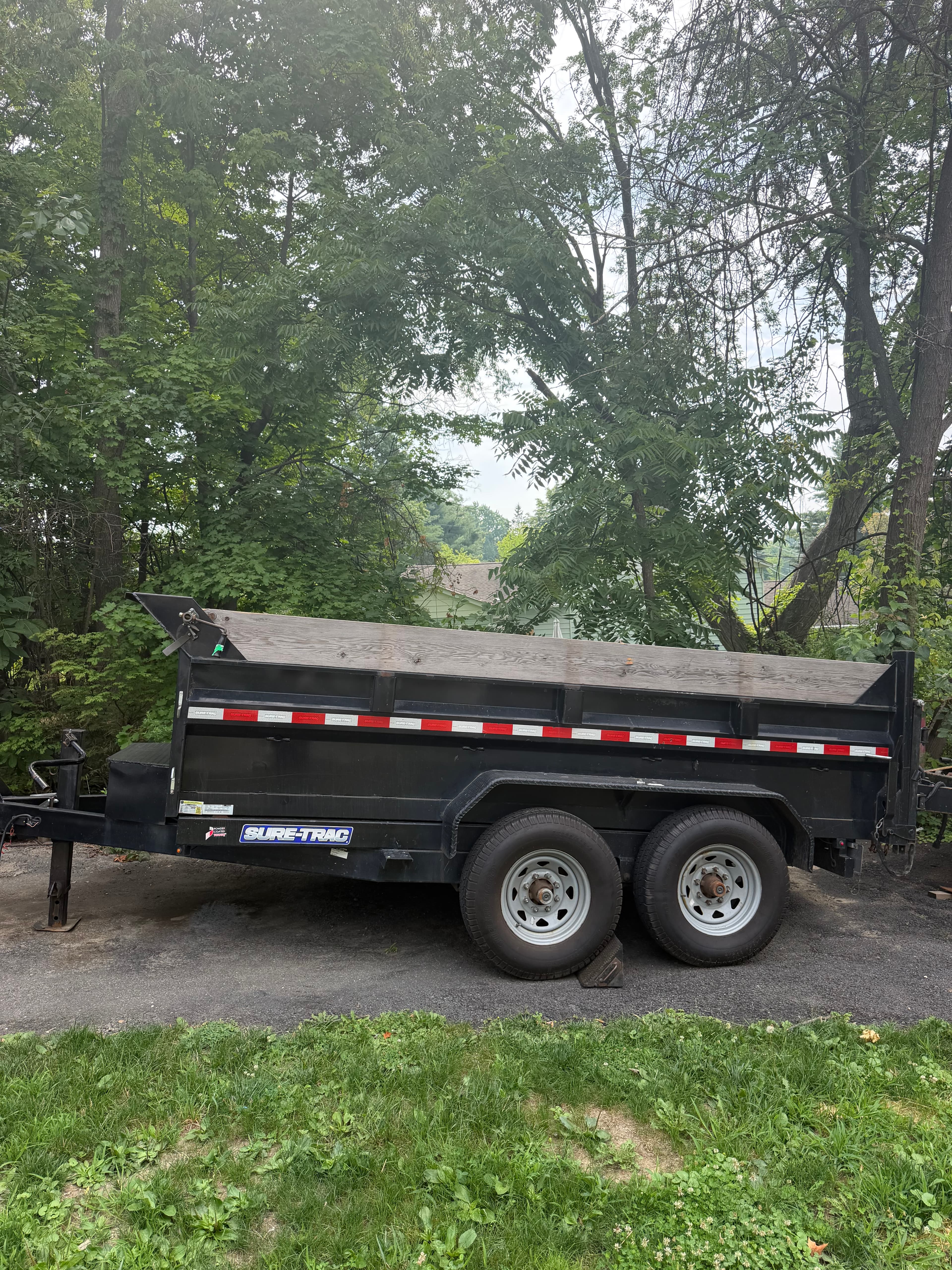 Black Sure-Trac dump trailer with wooden sideboards parked on a driveway near green trees.